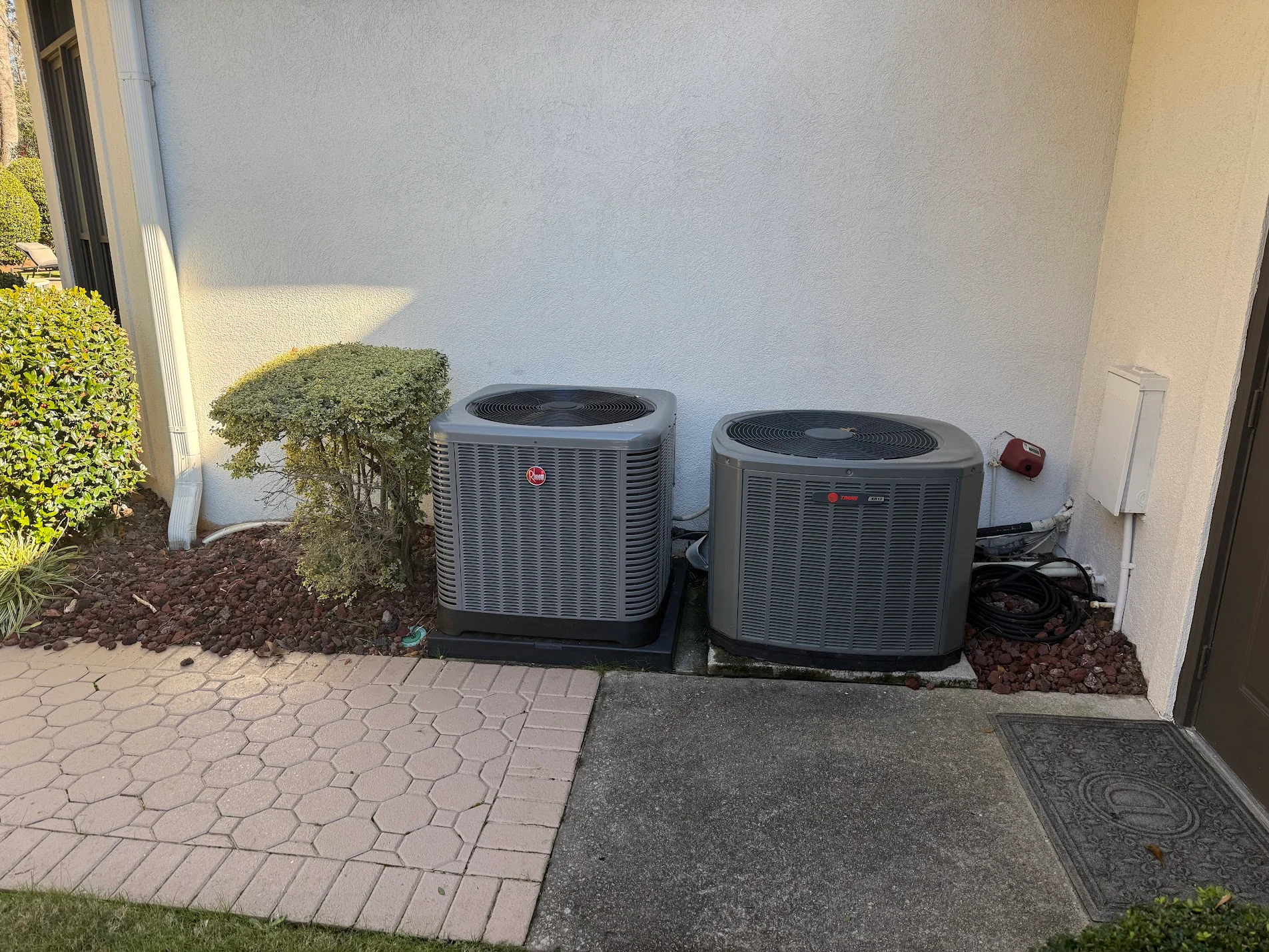 Two gray AC condenser units with black fan tops installed on concrete pads with rock landscaping
