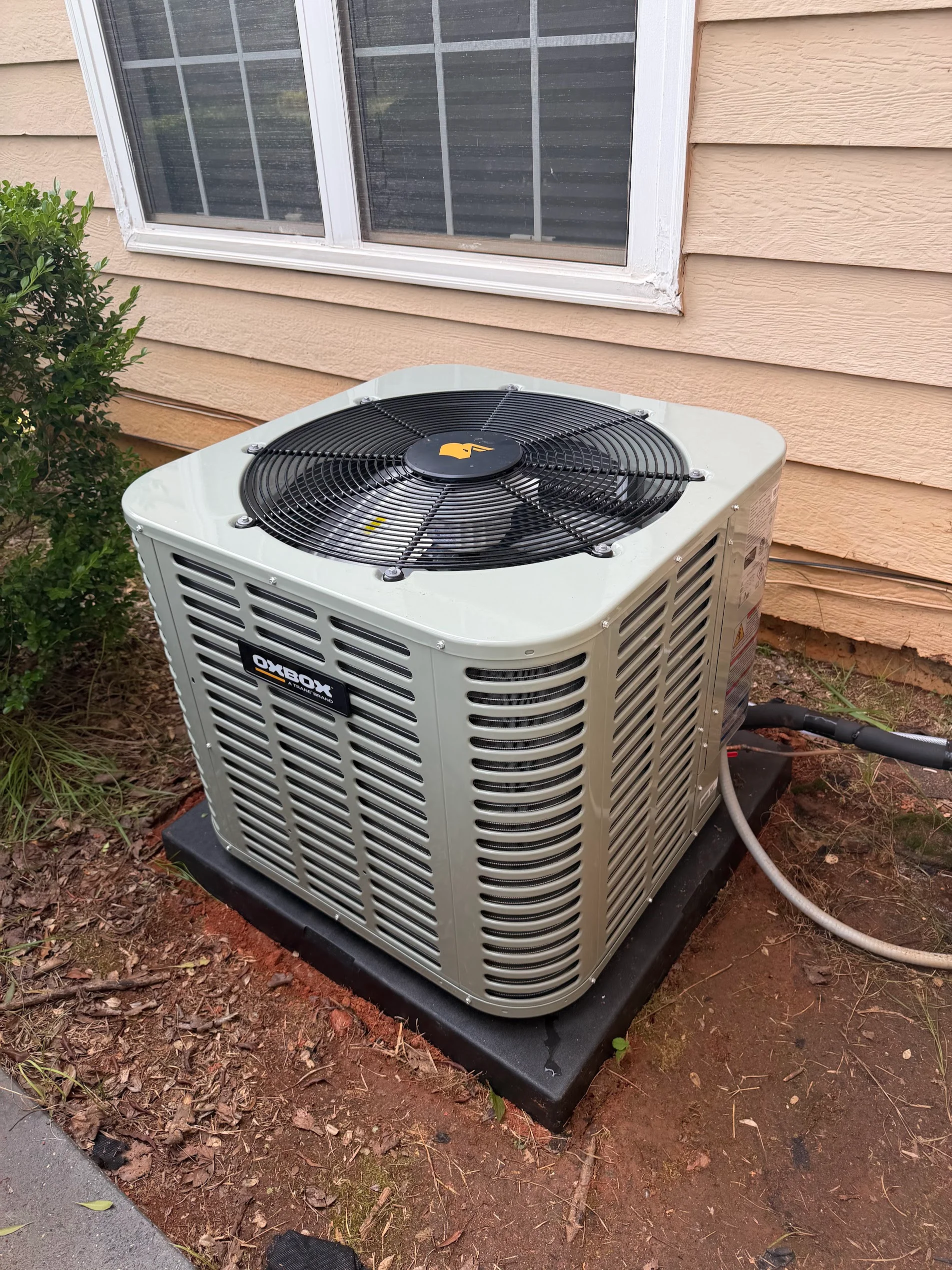 Beige air conditioning condenser unit with black fan motor installed on concrete pad against tan siding