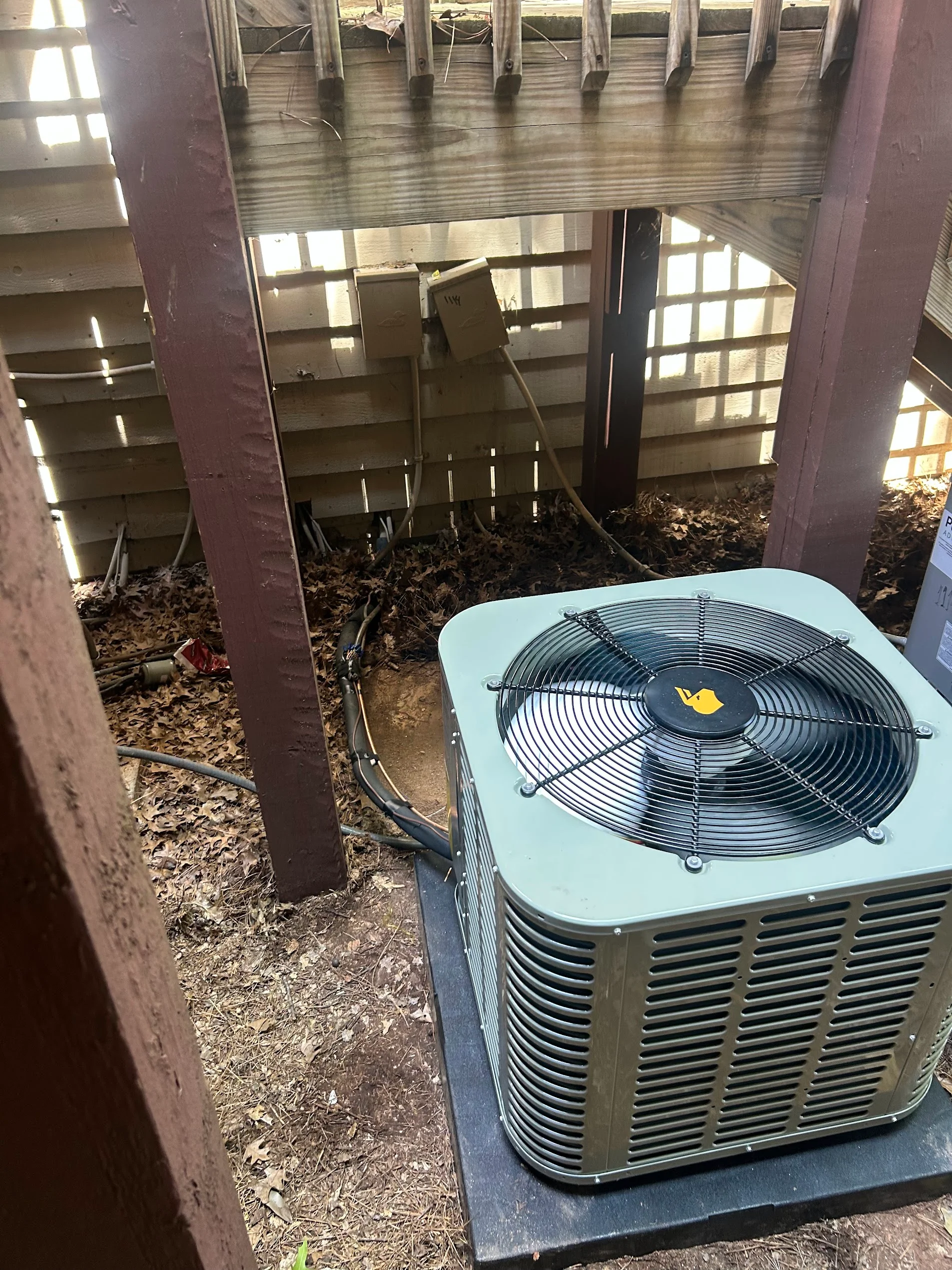 Light blue AC condenser unit with fan mounted on concrete pad under wooden structure with support posts