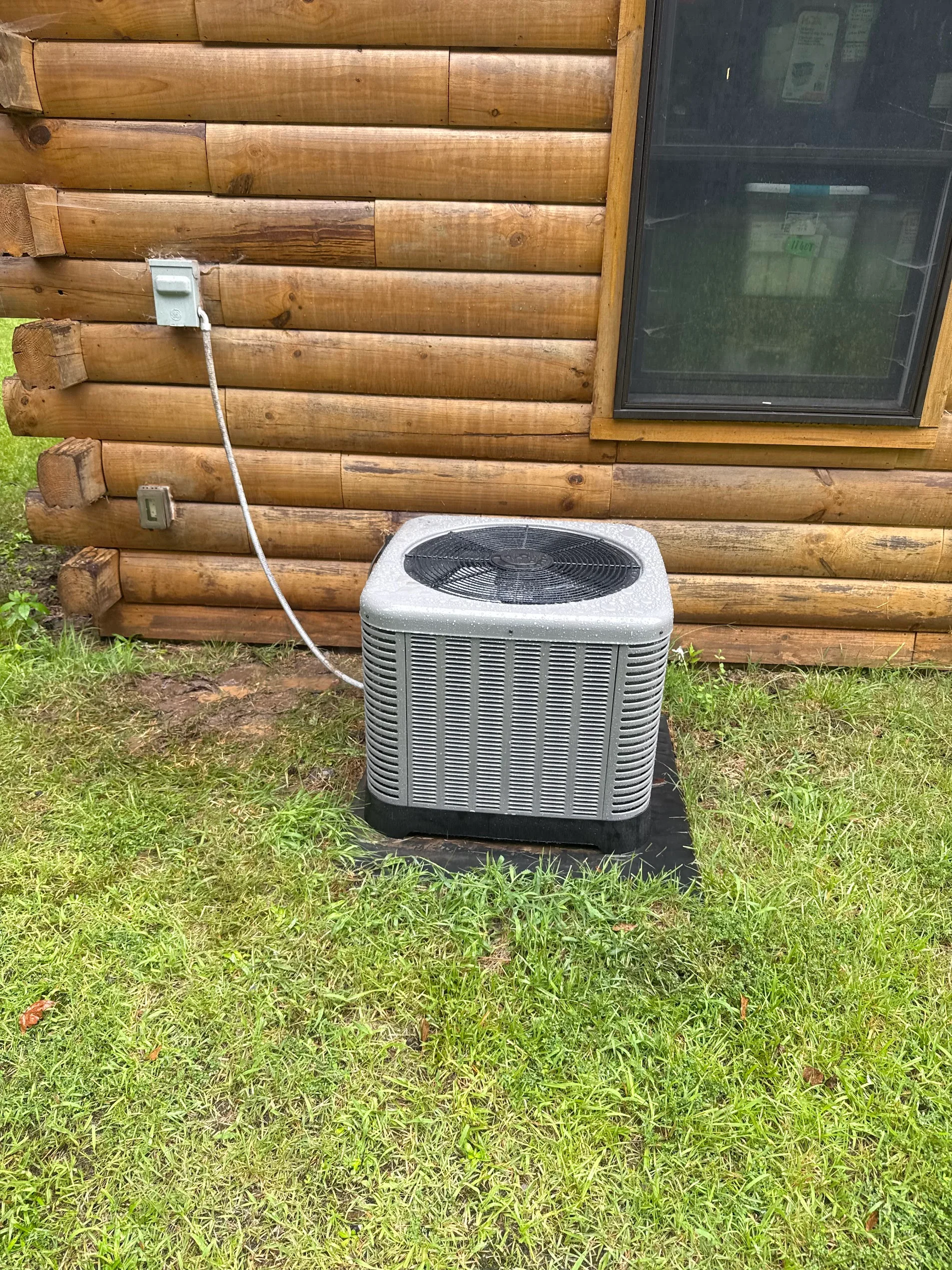 New AC condenser unit installed on concrete pad beside log cabin with electrical line connection