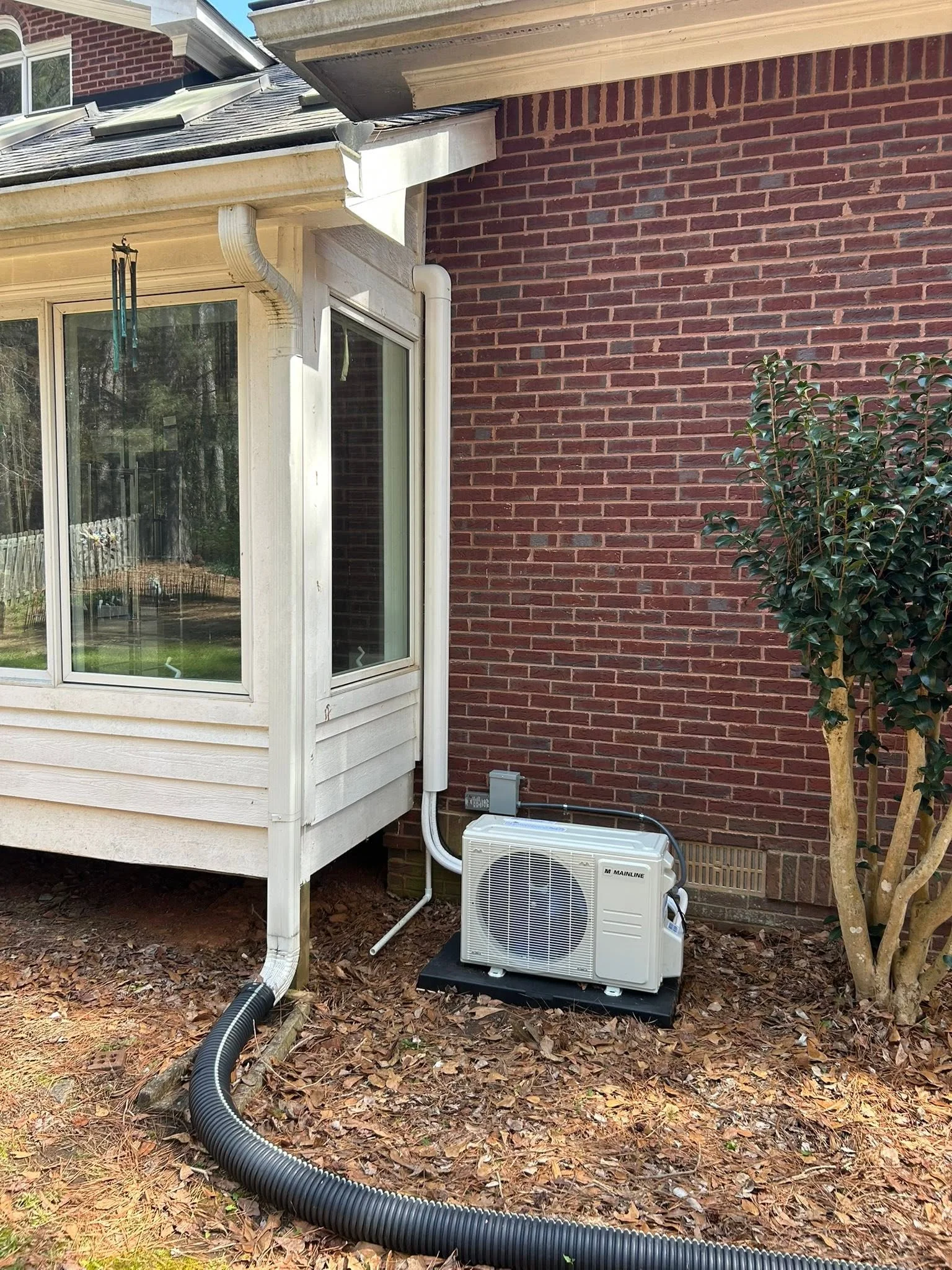 White mini-split heat pump outdoor unit installed on concrete pad with refrigerant lines and drainage hose routed to brick home