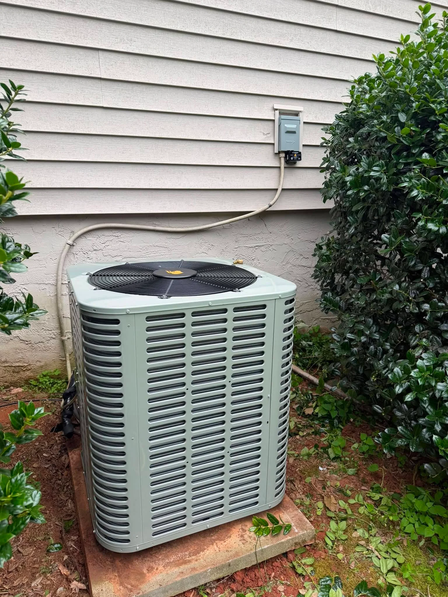 Light-green heat pump outdoor unit on concrete pad with disconnect switch and refrigerant lines against white horizontal siding