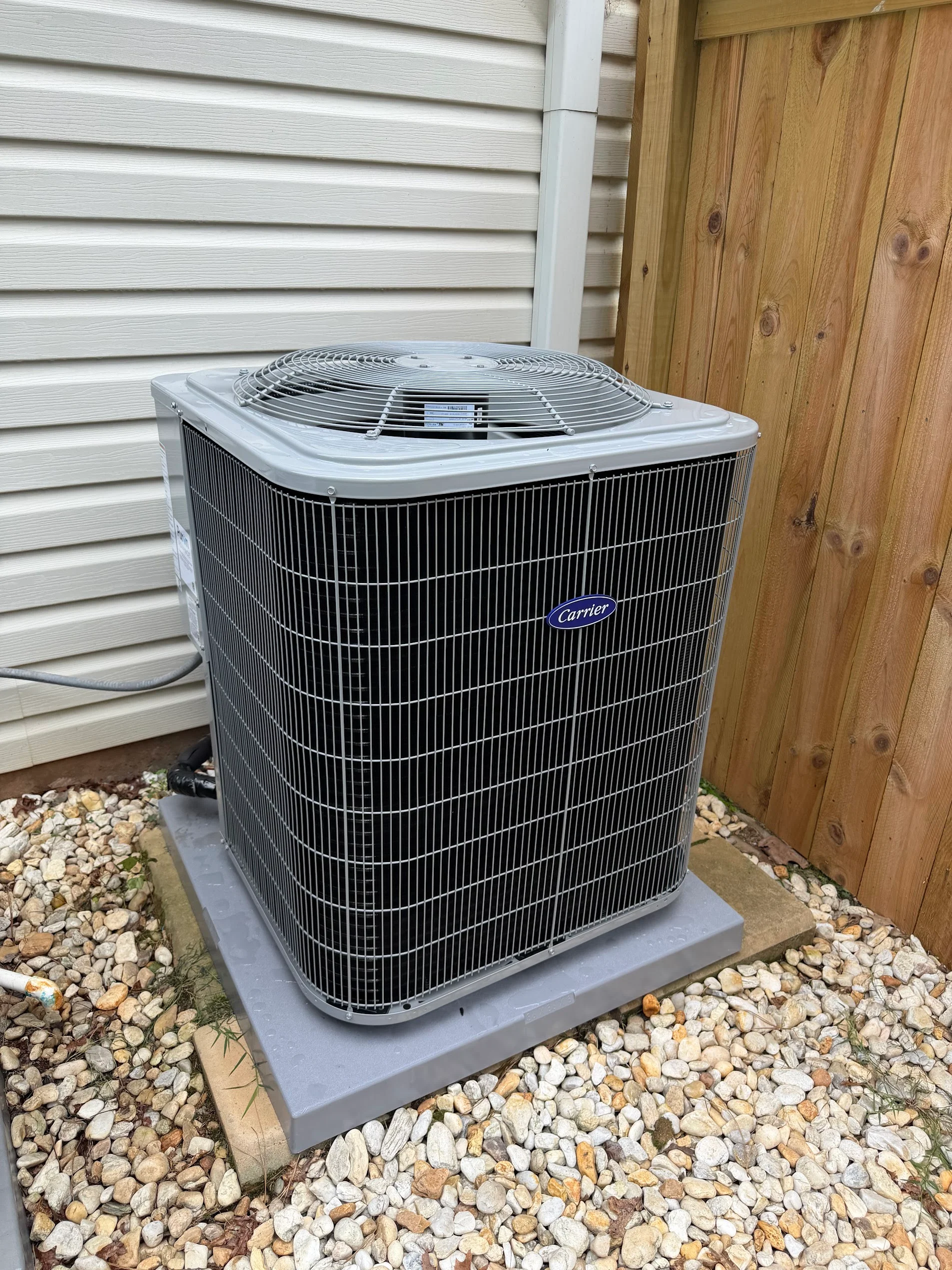 Carrier AC condenser unit on concrete pad with gravel base, white vinyl siding backdrop